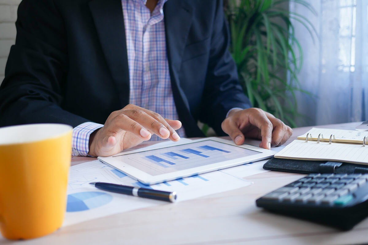Businessperson analyzing bar charts on a tablet at a desk with a calculator, pen, yellow coffee mug, and notebook.